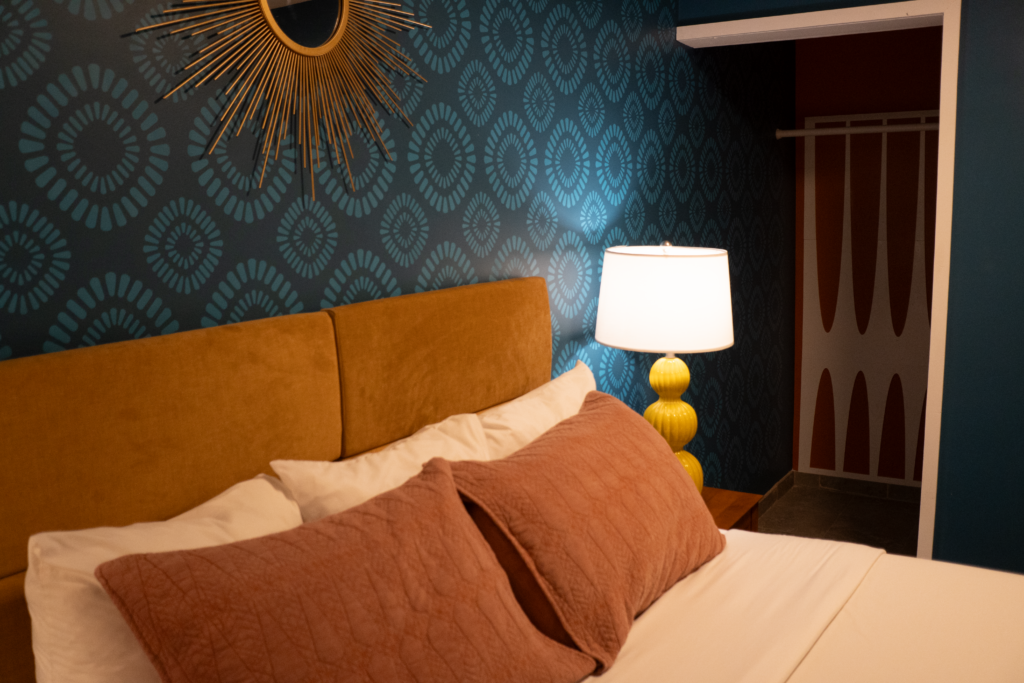 A close-up of a bed with a brown headboard, white bedding, and red pillows, with a yellow lamp and blue wallpaper in the background.