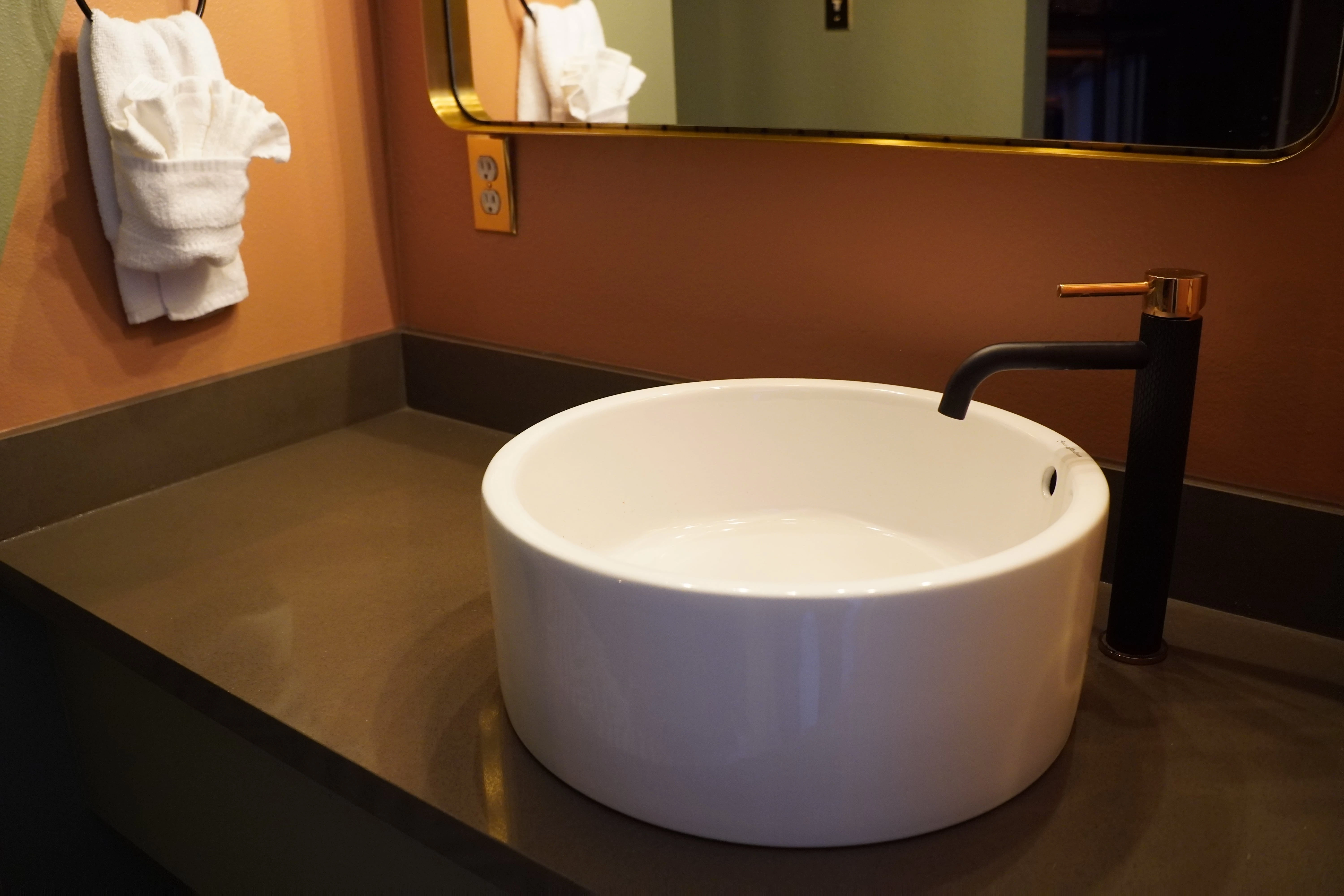 A modern bathroom sink with a white bowl, black faucet, and gold mirror frame, with peach walls and a gray countertop.