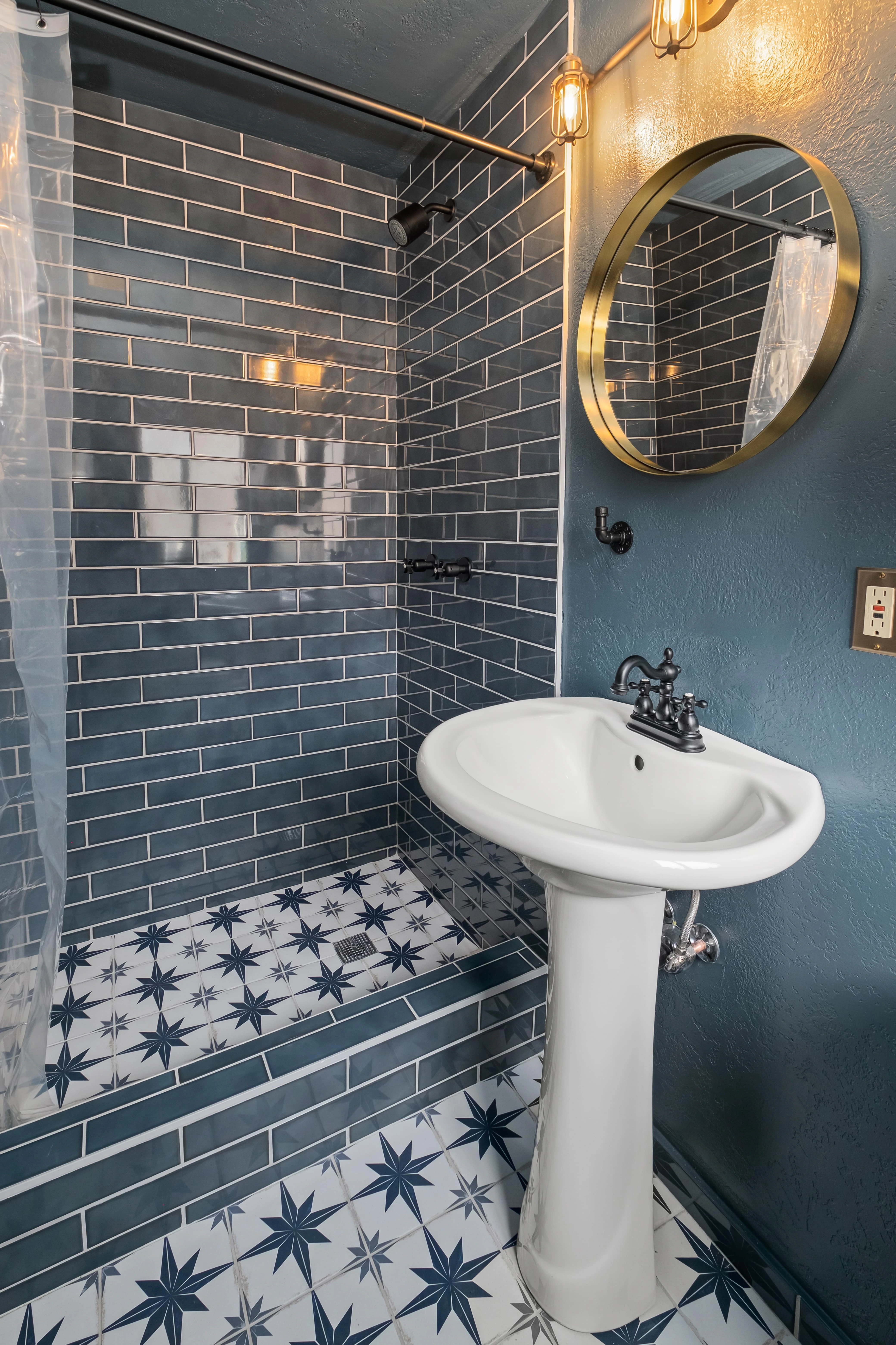 A clean and stylish bathroom with a tiled shower, pedestal sink, and round mirror.