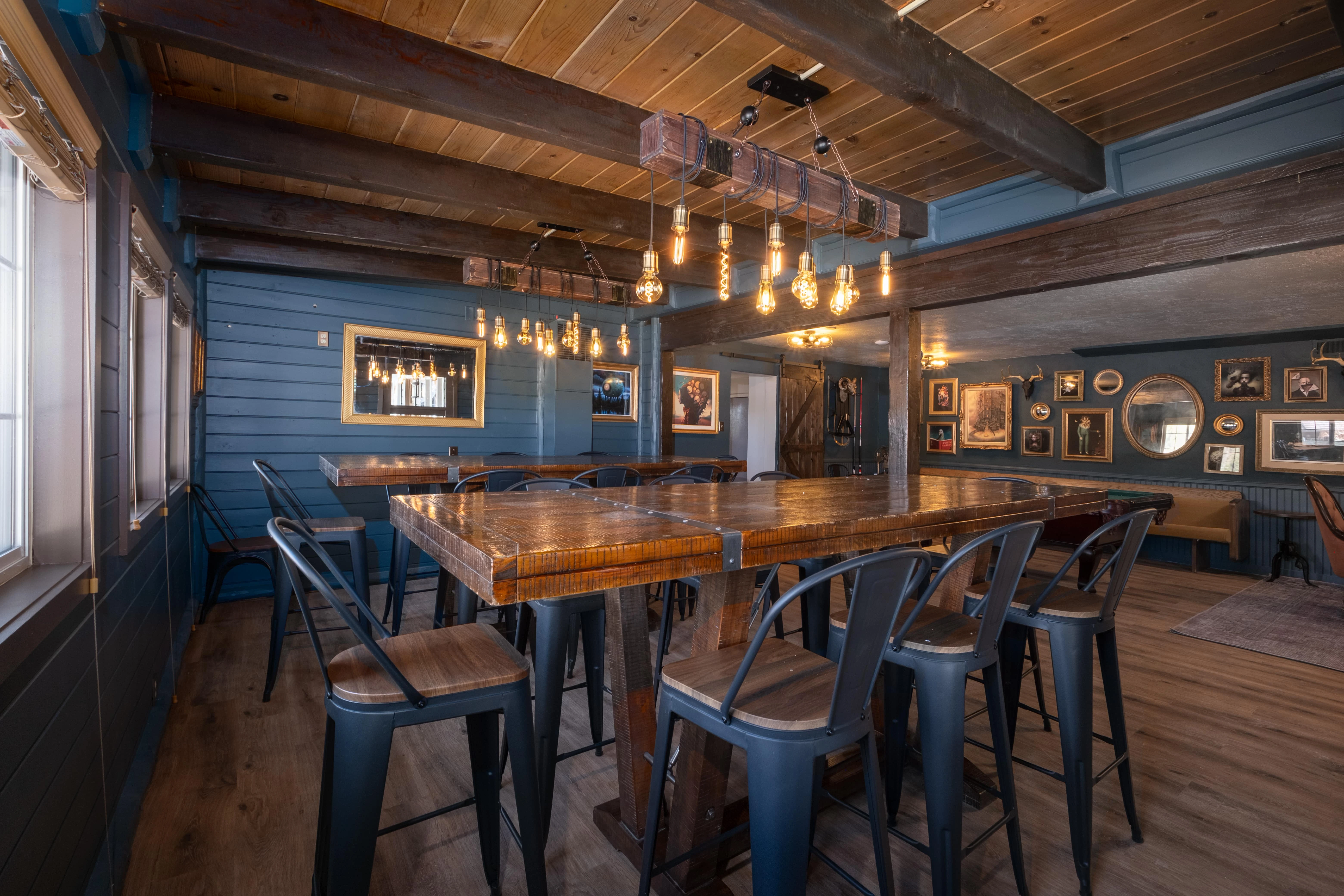 A bar area with a wooden table and metal chairs, blue walls, exposed beams, and hanging lights.