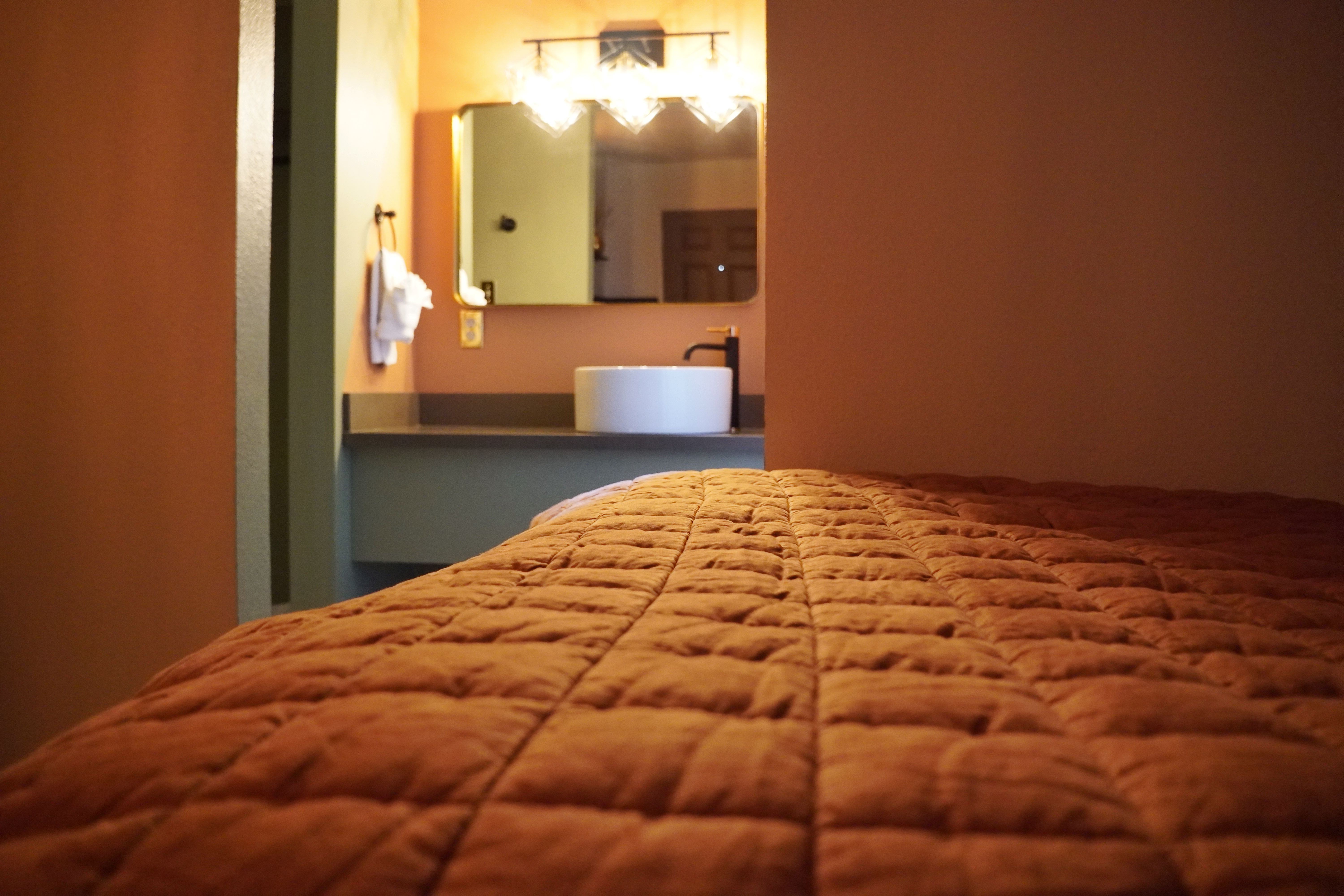 A bed with an orange comforter in the foreground and a bathroom with a sink and mirror in the background.