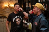 Two men drinking at a bar with liquor bottles in the background.