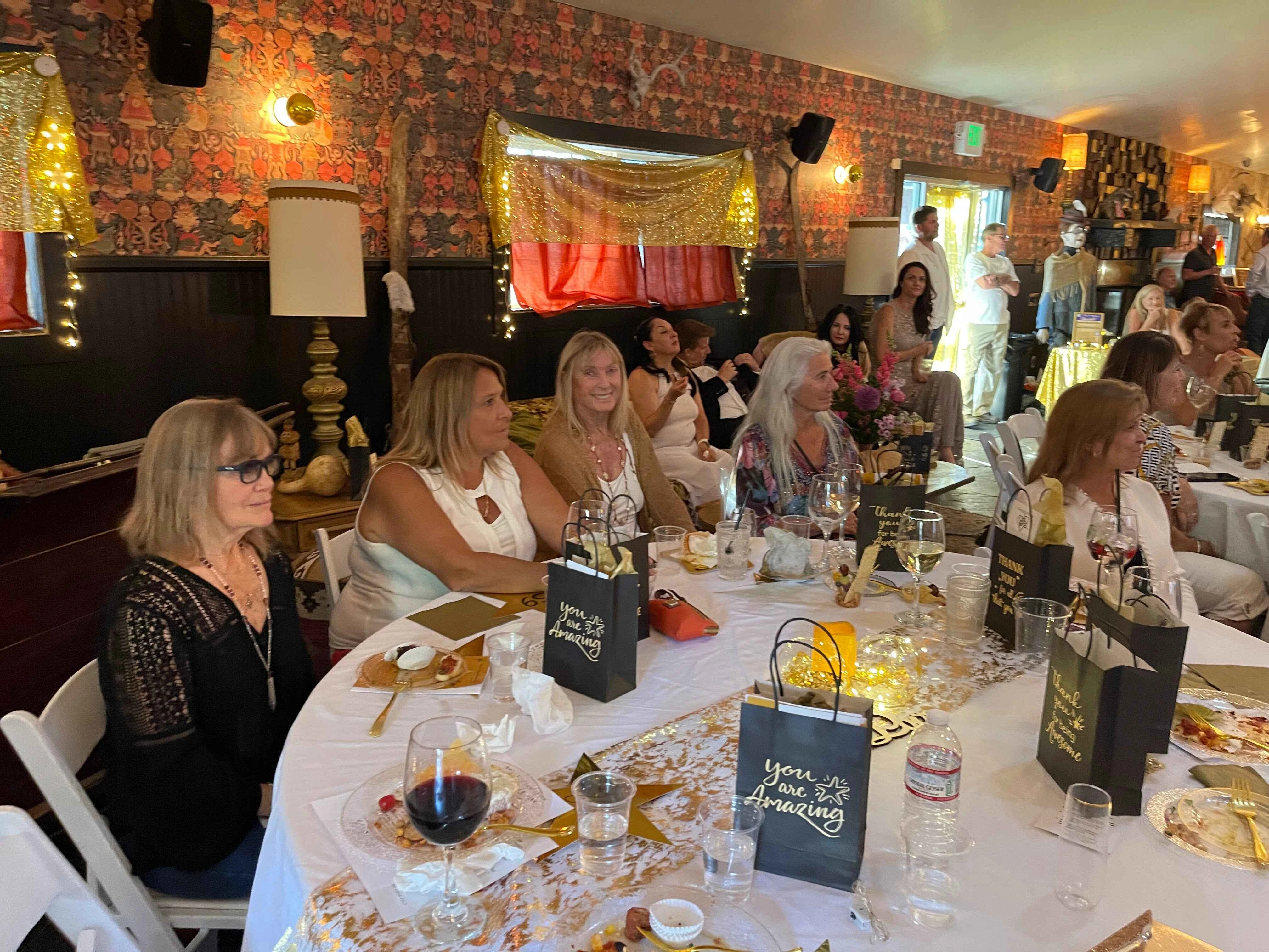 A group of people sitting at a round table with a white tablecloth, wine glasses, and gift bags.