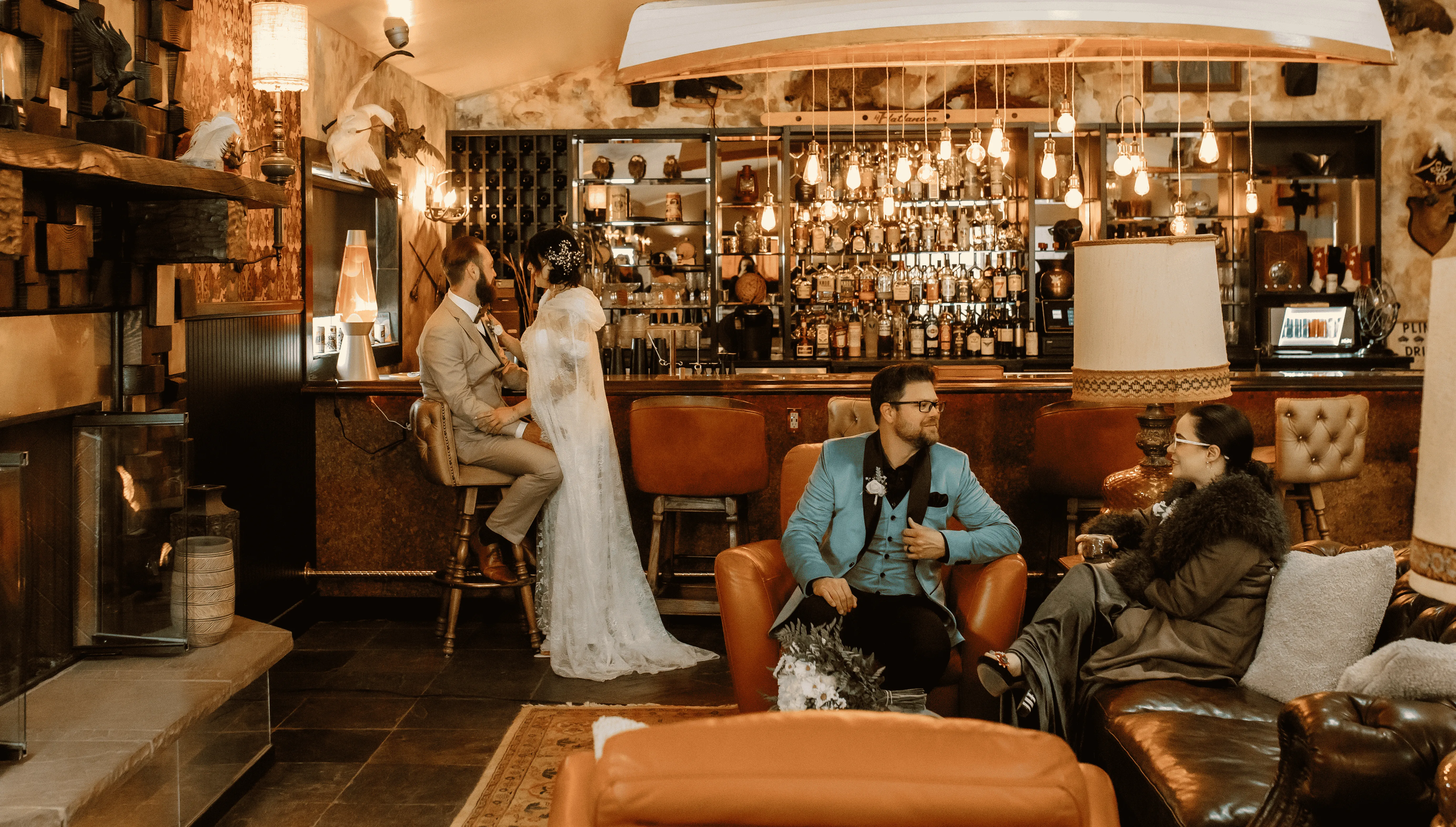 A dimly lit hotel bar with a fireplace, leather armchairs, and a wooden bar with people sitting and socializing.