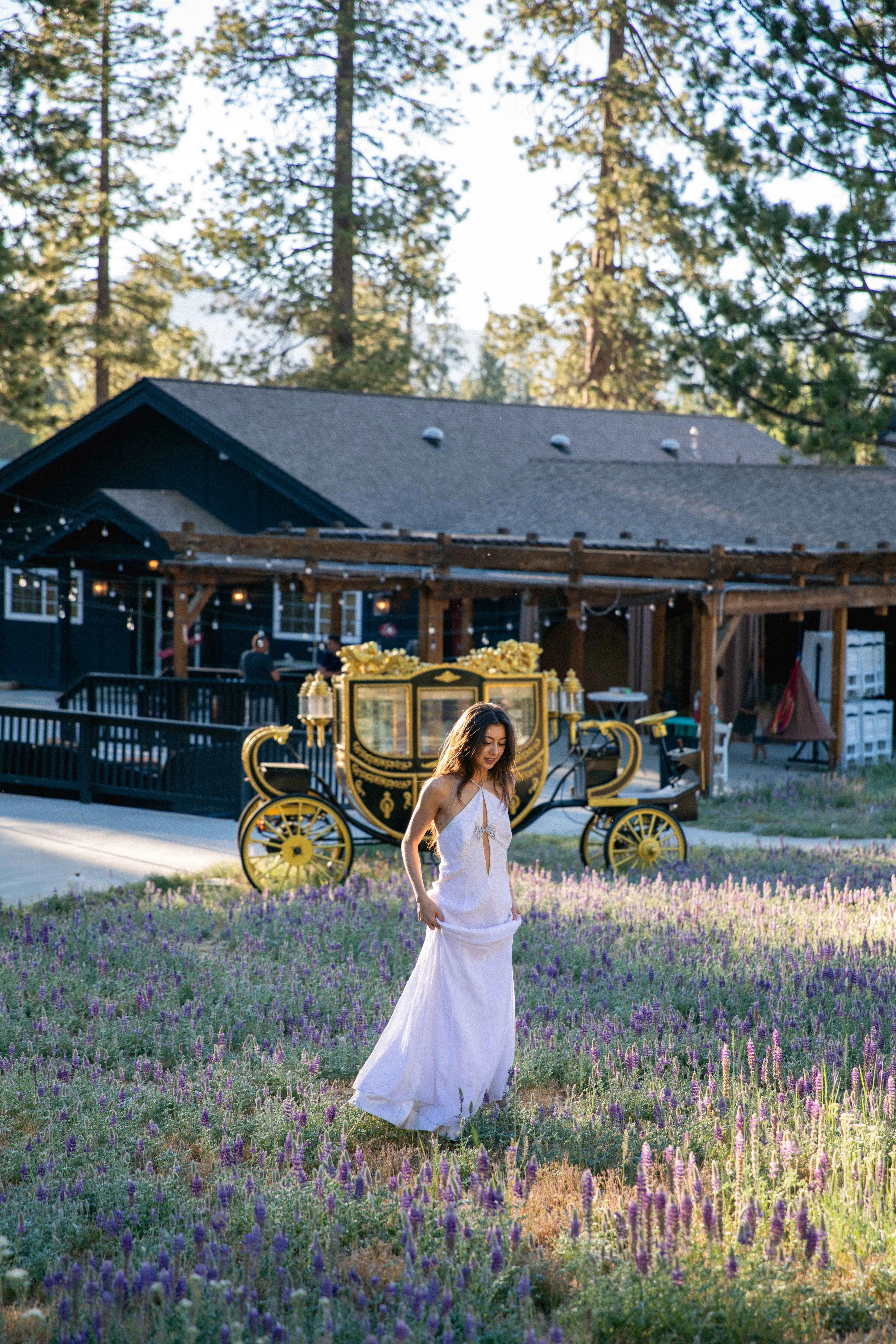 A woman in a white dress standing in a field of flowers with a building and carriage in the background.