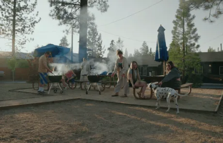 A group of people gathered around grills and outdoor furniture in a dirt area with a building in the background.
