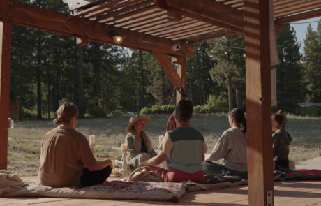 A group of people sitting on cushions under a wooden pergola in a forested area.